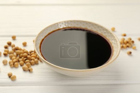 Tasty soy sauce in bowl and soybeans on white wooden table, closeup