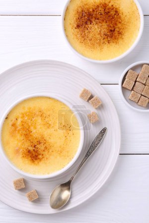 Foto de Deliciosa crema brulee en cuencos, terrones de azúcar y cuchara en mesa de madera blanca, puesta plana - Imagen libre de derechos