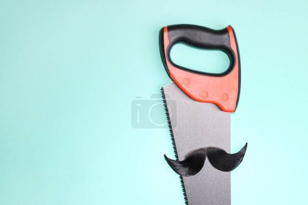 Téléchargez les photos : Visage d'homme fait de moustache artificielle et scie à main sur fond bleu clair, vue de dessus. Espace pour le texte - en image libre de droit