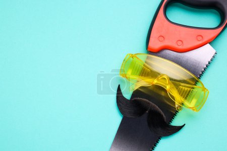 Téléchargez les photos : Visage d'homme fait de moustache artificielle, lunettes de sécurité et scie à main sur fond bleu clair, vue de dessus. Espace pour le texte - en image libre de droit