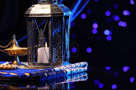 Arabic lantern, misbaha, Aladdin magic lamp and folded prayer mat on mirror surface against blurred lights at night. Space for text