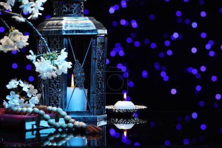 Arabic lantern, Quran, misbaha, burning candle and flowers on mirror surface against blurred lights at night. Space for text
