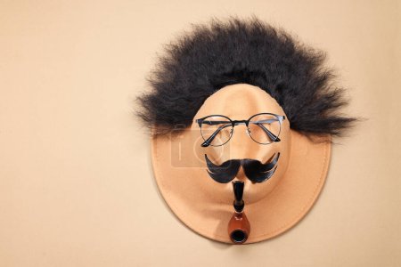Téléchargez les photos : Visage d'homme en cheveux artificiels, moustache, lunettes et chapeau sur fond beige, vue de dessus. Espace pour le texte - en image libre de droit