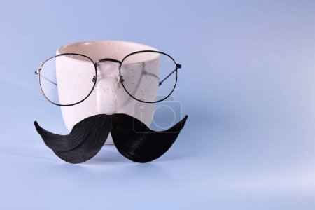 Téléchargez les photos : Visage d'homme fait de moustache artificielle, lunettes et tasse sur fond bleu clair. Espace pour le texte - en image libre de droit