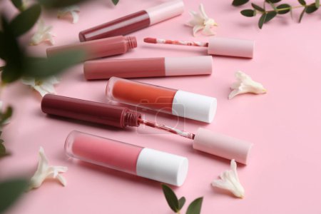 Foto de Brillos labiales diferentes, aplicadores, flores y hojas verdes sobre fondo rosa - Imagen libre de derechos