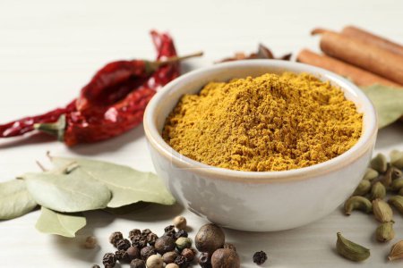 Curry powder in bowl and other spices on white wooden table, closeup