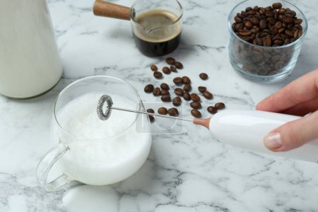 Woman whisking milk in cup with mini mixer (milk frother) at white marble table, closeup