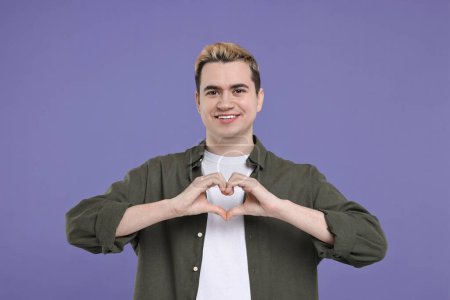 Téléchargez les photos : Jeune homme montrant geste de coeur avec les mains sur fond violet - en image libre de droit