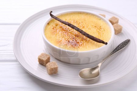 Téléchargez les photos : Délicieuse crème brulée dans un bol, gousse de vanille, cubes de sucre et cuillère sur table en bois blanc, gros plan - en image libre de droit