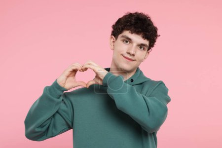 Téléchargez les photos : Jeune homme heureux montrant geste de coeur avec les mains sur fond rose - en image libre de droit