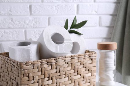 Téléchargez les photos : Rouleaux de papier toilette dans le panier en osier, décor floral et tampons de coton sur la table près du mur de briques blanches - en image libre de droit
