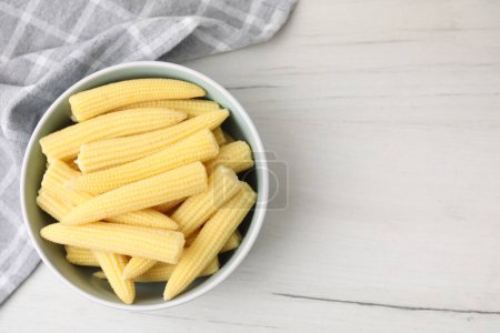 Tasty fresh yellow baby corns in bowl on white wooden table, top view. Space for text