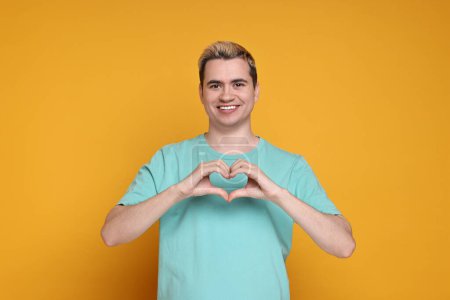 Téléchargez les photos : Jeune homme montrant un geste de coeur avec les mains sur fond orange - en image libre de droit