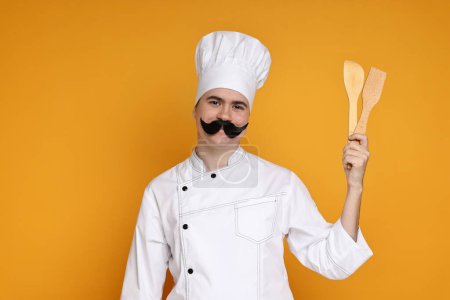 Portrait of happy confectioner with funny artificial moustache holding wooden spatulas on orange background