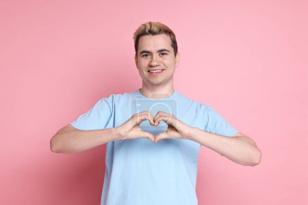 Téléchargez les photos : Jeune homme montrant geste de coeur avec les mains sur fond rose - en image libre de droit