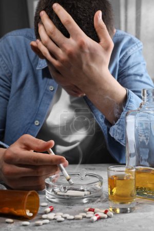 Alcohol and drug addiction. Man with smoldering cigarettes, whiskey and pills at grey textured table