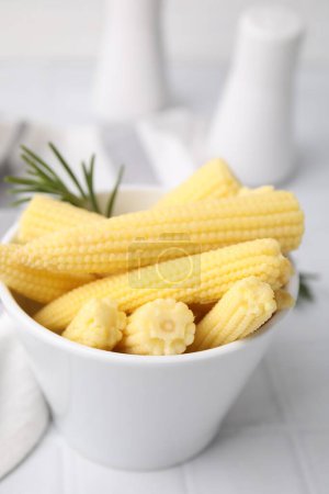 Tasty fresh yellow baby corns in bowl on white tiled table, closeup