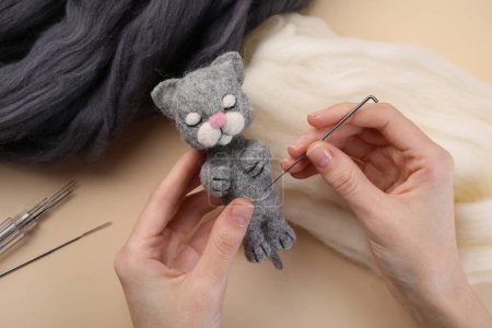 Woman felting cute toy cat from wool at beige table, closeup