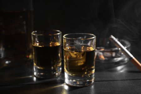 Alcohol addiction. Whiskey in glasses, smoldering cigarette and ashtray on black wooden table