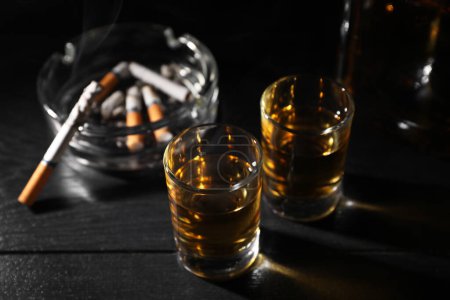 Téléchargez les photos : L'alcoolisme. Whisky dans les verres, cigarette fumante et cendrier sur table en bois noir, gros plan - en image libre de droit