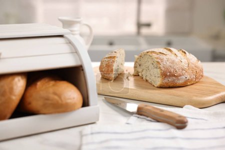 Wooden bread basket with freshly baked loaves and knife on white marble table in kitchen