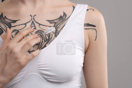 Frau trägt heilende Creme auf ihre Tattoos vor grauem Hintergrund auf, Nahaufnahme
