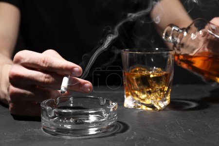 Téléchargez les photos : L'alcoolisme. Homme avec cigarette fumante versant du whisky dans du verre à une table sombre texturée, gros plan - en image libre de droit
