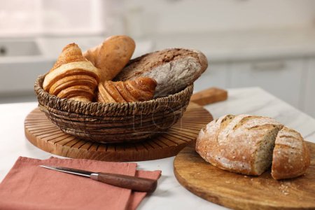 Photo for Wicker bread basket with freshly baked loaves and knife on white marble table in kitchen - Royalty Free Image