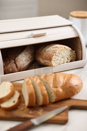 Brotkorb aus Holz mit frisch gebackenen Broten und Messer auf weißem Marmortisch in der Küche