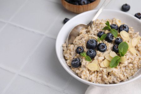 Tasty oatmeal with blueberries, mint and almond flakes in bowl on white tiled table, closeup. Space for text