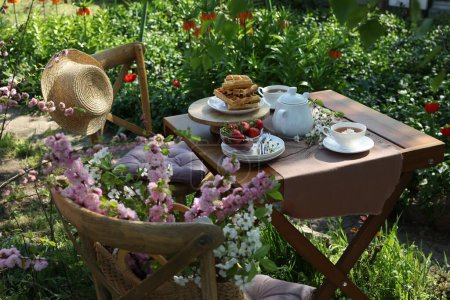Téléchargez les photos : Belles fleurs de printemps, gaufres fraîchement cuites et fraises mûres sur la table servies pour boire le thé dans le jardin - en image libre de droit
