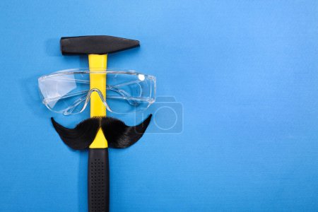 Téléchargez les photos : Visage d'homme fait de moustache artificielle, lunettes de sécurité et marteau sur fond bleu, vue de dessus. Espace pour le texte - en image libre de droit