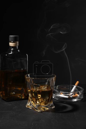 Adicción al alcohol. Whisky, cigarrillos humeantes y cenicero en mesa de textura oscura