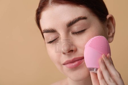 Téléchargez les photos : Se laver le visage. Jeune femme avec brosse nettoyante sur fond beige, gros plan - en image libre de droit