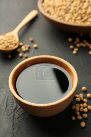 Leckere Sojasauce in Schüssel und Sojabohnen auf schwarzem Tisch, Nahaufnahme