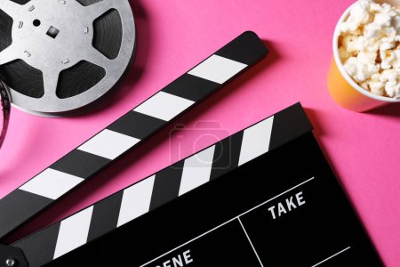 Clapperboard, popcorn and film reel on pink background, flat lay