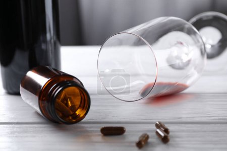 Téléchargez les photos : Alcool et toxicomanie. Verre renversé avec vin rouge, bouteille et pilules sur table en bois blanc, gros plan - en image libre de droit