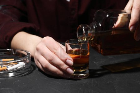Adicción al alcohol. Mujer vertiendo whisky de botella en vidrio en la mesa de textura oscura, primer plano