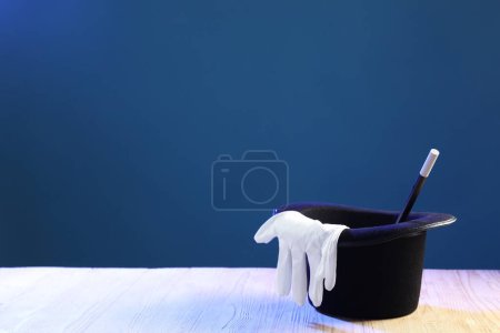 Magician's hat, wand and gloves on wooden table against blue background, space for text