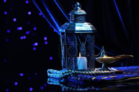 Arabic lantern, misbaha, Aladdin magic lamp and folded prayer mat on mirror surface against blurred lights at night. Space for text
