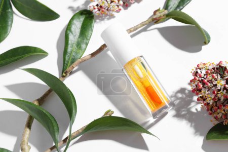 Orange lip gloss, branch, green leaves and flowers on white background, above view