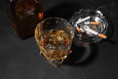 Adicción al alcohol. Whisky con cubitos de hielo, cigarrillos humeantes y cenicero en mesa de textura oscura, por encima de la vista