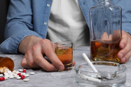 Alcohol and drug addiction. Man with whiskey, pills and smoldering cigarettes at grey textured table, closeup