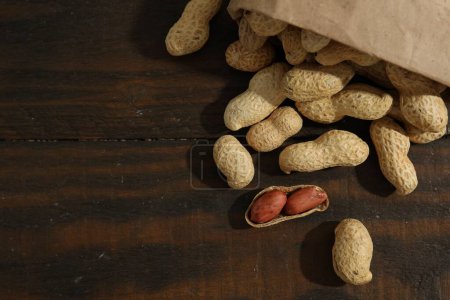 Téléchargez les photos : Paper bag with fresh unpeeled peanuts on wooden table, top view. Space for text - en image libre de droit