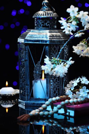 Arabic lantern, Quran, misbaha, burning candle and flowers on mirror surface against blurred lights at night