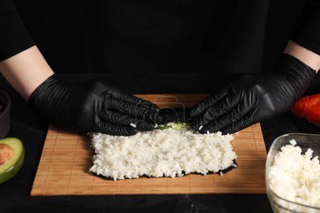 Téléchargez les photos : Chef en gants enveloppant un rouleau de sushi à table noire, gros plan - en image libre de droit
