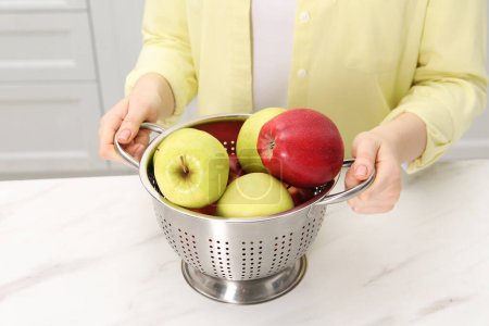 Woman holding colander with fresh apples at white marble table in kitchen, closeup