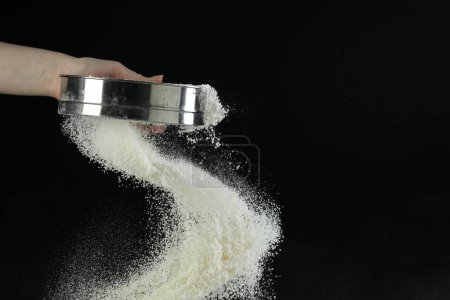 Woman sieving flour against black background, closeup. Space for text