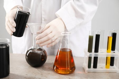 Woman pouring black crude oil from beaker into flask at grey table against light background, closeup