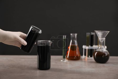 Téléchargez les photos : Femme versant du pétrole brut noir dans un bécher à table grise sur fond sombre, gros plan - en image libre de droit
